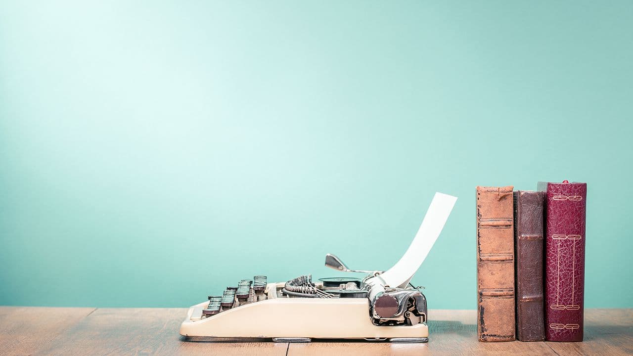 Retro classic typewriter from circa 1950s with sheet of paper and aged books on wooden desk front aquamarine wall background. Nostalgic writer's work place concept. Vintage old style filtered photo | ©BrAt82 - stock.adobe.com 