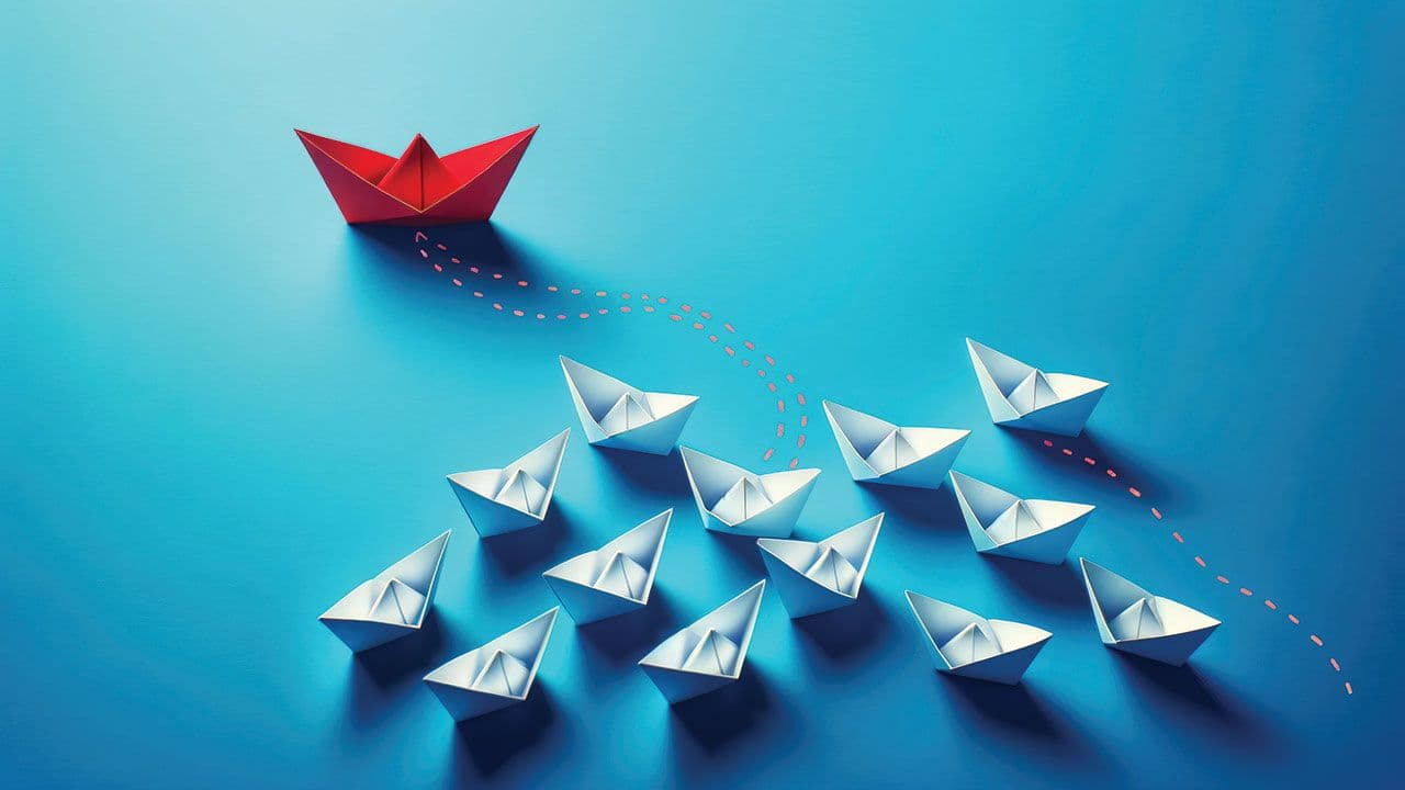 Leadership and innovation, featuring a single red paper boat forging a unique path ahead of a fleet of white paper boats on a striking blue background. | Image Credit: ©John - stock.adobe.com