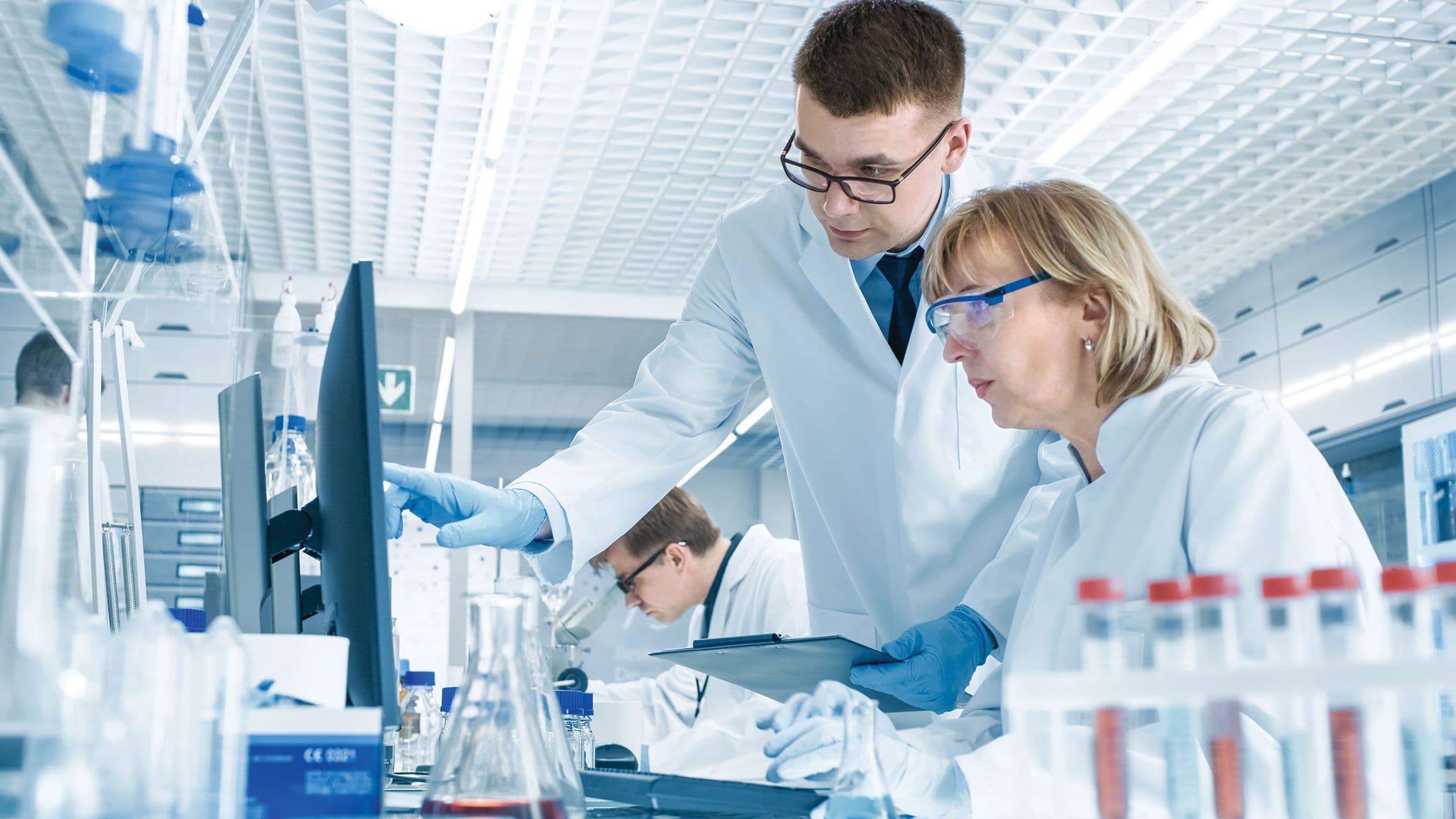 In Modern Laboratory Senior Female Scientist Has Discussion with Young Male Laboratory Assistant. He Shows Her Data Charts on a Clipboard, She Analyzes it and Enters It into Her Computer. | Image Credit: © Gorodenkoff - stock.adobe.com