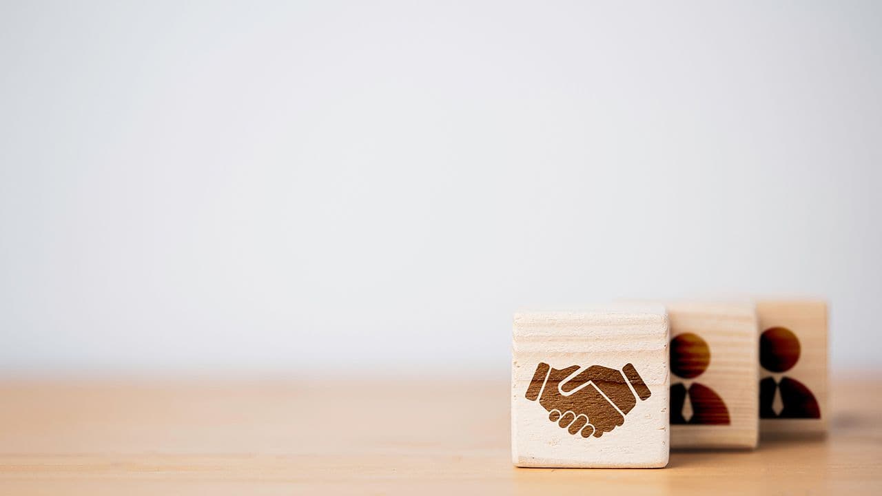 Hand shaking which print screen on wooden cube block in front of human icon for business deal and agreement concept. | Image Credit: © Dilok - stock.adobe.com