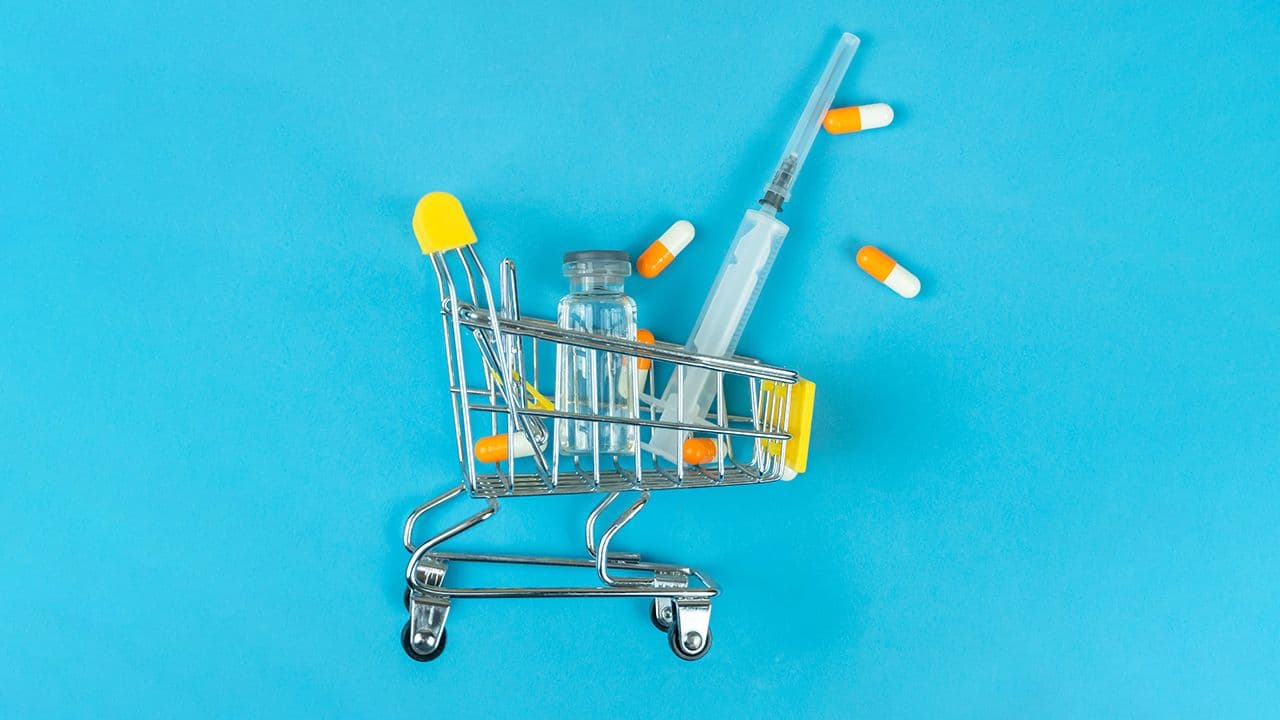 Shopping cart with medical ampoule, syringe and capsules on a blue background, copy space, top view. Concept of purchasing medicines, increasing drug prices. Vertical photo | Image Credit: © yaroslav1986 - Stock.adobe.com